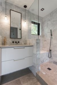 A bathroom with marble walls and floors, two sinks, mirror, shower head and window.